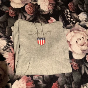 Grå hoodieklänning från GANT - Säljer en stilren grå hoodieklänning från GANT med ett broderat emblem på bröstet. Klänningen har långa ärmar och en praktisk magficka. Perfekt för en avslappnad stil.