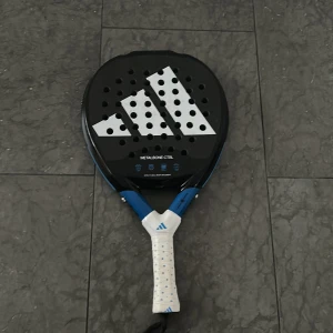 Padelracket Adidas Metalbone CTRL - Säljer ett Adidas Metalbone CTRL padelracket. Racketen har en svart och vit design med blå detaljer och ett perforerat mönster. Handtaget är lindat med ett vitt grepp med blå logotyp. Perfekt för kontrollspelare som söker precision och balans. Har en liten spricka men inga andra problem. 