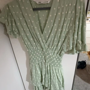 Grön prickig klänning från Zara - Säljer en söt grön klänning från Zara med vita prickar och v-ringad design. Klänningen har korta ärmar och en smockad midja som ger en fin passform. Perfekt för vår och sommar!