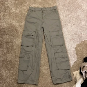 Grå cargobyxor från H&M Divided - Snygga grå cargobyxor från H&M Divided med flera praktiska fickor. Byxorna har en loose passform och är perfekta för en avslappnad stil. De har knappar och dragkedja i midjan.