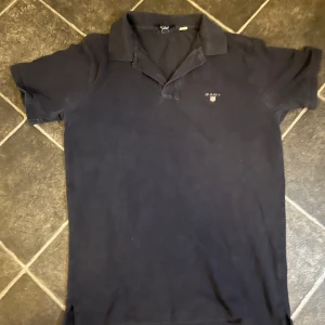 Blå  pikétröja från GANT - Snygg svart pikétröja från GANT med klassisk krage och korta ärmar. Perfekt för en stilren och avslappnad look. Nypris 1000kr, mitt pris 250kr, storleken är M men passar S.