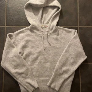 Grå hoodie från Gina Tricot - Mysig grå hoodie från Gina Tricot med justerbar dragsko i huvan. Perfekt för kyliga dagar med sin mjuka och bekväma passform. Ribbstickade muddar vid ärmslut och nederkant ger extra komfort.