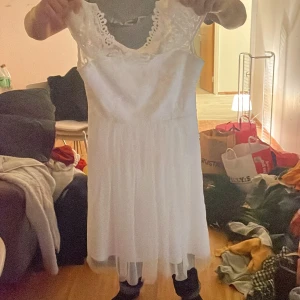 Vit klänning med spetsdetaljer - Elegant vit klänning med vackra spetsdetaljer på överdelen och en luftig kjol. Klänningen har en v-ringad design och är ärmlös, vilket ger en romantisk och stilren look.