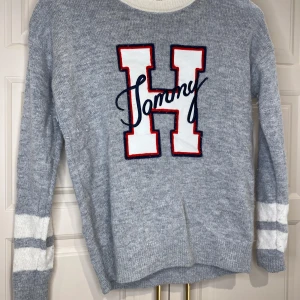 Grå tröja från Tommy Hilfiger - Säljer en snygg grå tröja från Tommy Hilfiger med ett stort 'H' och 'Tommy' broderat på framsidan. Tröjan har vita detaljer vid halsringning och ärmslut samt långa ärmar. Perfekt för en avslappnad stil.