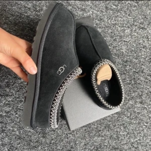 Svarta tofflor från UGG - Mysiga svarta tofflor från UGG i mocka med en mjuk insida. De har en dekorativ stickad kant i grått och vitt som ger en stilren look. Perfekta för att hålla fötterna varma under kyliga dagar. rengöra mer innan jag skickar.