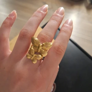 Guldfärgad ring med fjärilar - Elegant guldfärgad ring prydd med två detaljerade fjärilar. Perfekt för att ge en touch av naturinspirerad skönhet till din stil. Ringen har en unik design som fångar ljuset vackert.