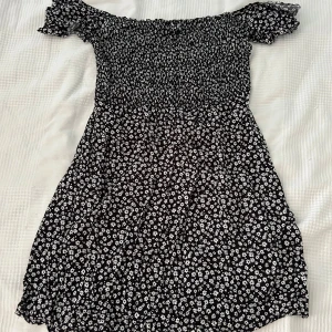 Svart blommig klänning från FB Sister - Säljer en superfin svart klänning med vita blommor från FB Sister. Den har en offshoulder-design och korta ärmar med volangdetaljer 🌸
