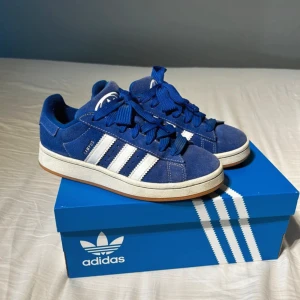 Blå Adidas sneakers - Säljer ett par snygga blå Adidas sneakers i modellen Adidas campus 00’s. Skorna är blåa med vita detaljer och har en klassisk design med snörning. De kommer i originalkartong samt andra skosnören och är i bra skick, men använd ett par gånger