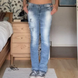 Blå jeans med slitningar - Säljer ett par snygga blå jeans med coola slitningar och en unik patch-detalj på låret. De har en låg midja och är uppsprättade vid fötterna. Passar också till någon som är lite längre. Jag är 178 cm❤️Storlek 36 enligt etiketten men passar även 38.