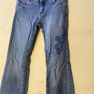 Blå jeans med broderi - Säljer ett par blå jeans med snyggt blommigt broderi på ena benet. De har en klassisk femficksdesign och är i en straight fit. Tyvärr finns det en reva vid bakfickan som kan behöva lagas. Perfekta för en avslappnad stil! Storlek 146