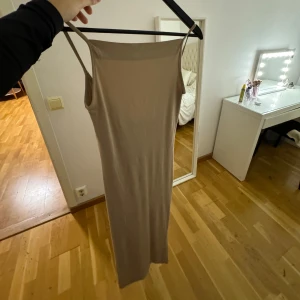Gina tricot klänning  - Aldrig använt köpt för 449kr Säljer en stilren beige långklänning med smala axelband. Klänningen är ärmlös och har en enkel, elegant design som passar perfekt för både vardag och fest. Materialet ser ut att vara mjukt och bekvämt.