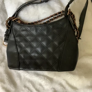 Svart quiltad axelväska med kedjedetaljer - Säljer en snygg svart axelväska i syntetmaterial med quiltat mönster. Väskan har coola kedjedetaljer i guld och en praktisk dragkedja på baksidan. Perfekt för både vardag och fest!