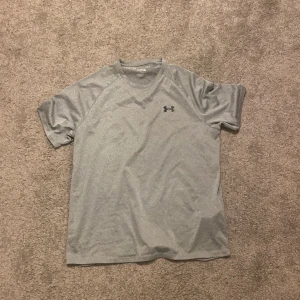 Grå t-shirt från Under Armour - Säljer en grå t-shirt från Under Armour. Den har en lös passform och är kortärmad, perfekt för träning. T-shirten är tillverkad i ett syntetmaterial som andas bra.