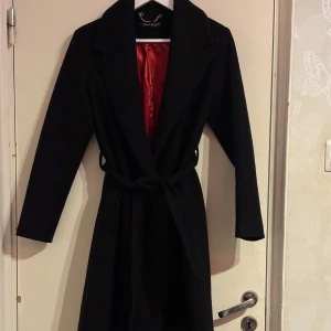 Svart kappa från Lord Mark - Elegant svart kappa från Lord Mark med röd insida. Den har ett bälte i midjan och långa ärmar, perfekt för en stilren look. Kappan är designad för att ge en klassisk och tidlös stil.