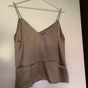 Beige satinlinne - Snyggt beige linne i satin. Linnet har smala axelband och en v-ringad design som ger en elegant look. Perfekt för att klä upp eller ner beroende på tillfälle. Finns vissa fläckar på linnet, ses på bilderna.