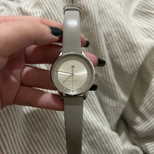 Grå klocka från Tommy Hilfiger - Snygg klocka från Tommy Hilfiger med en äkta grå läderrem och silverfärgad urtavla. Klockan har en design med märkets logotyp. Mindre “skada” på bandet men inget som syns när den är på! köpt för ca 1200kr, pris kan diskuteras:) 