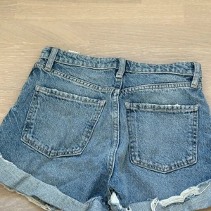 Blå jeansshorts från Zara - Snygga blå jeansshorts från Zara med slitna detaljer och fransig kant. De har en klassisk femficksdesign och knappar framtill. Perfekta för en avslappnad stil. Aldrig använda.
