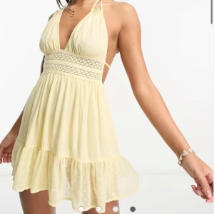Gul klänning från ASOS - Säljer en söt gul kort somrig klänning från ASOS med halterneck och spetsdetaljer i midjan och klänningen har öppen rygg. Klänningen har en volangkant. Den är helt ny 