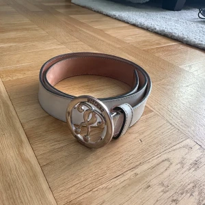 Vitt bälte med stor spänne från Moschino - Säljer ett stilrent vitt bälte från Moschino med ett stort, runt spänne i silverfärg. Bältet har en slät yta och flera justerbara hål för perfekt passform. Perfekt för att ge en lyxig touch till din outfit. Det finns en del slitage därför säljs den billigt. Köpt i Puerto Banos för 400 Euro 