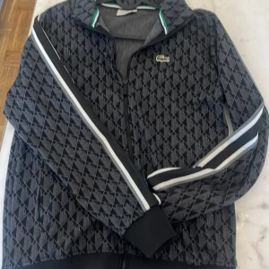 Mönstrad jacka från Lacoste - Snygg jacka från Lacoste med ett geometriskt mönster i svart och grått. Jackan har dragkedja framtill och långa ärmar med vita ränder längs sidorna. Den ikoniska Lacoste-loggan pryder bröstet. Perfekt för en stilren look!