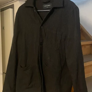 Svart overshirt från A Day's March - Snygg svart overshirt från A Day's March med knappar framtill. Perfekt för lager-på-lager-stil. Har en bröstficka och två sidofickor för extra funktionalitet. Passar bra till både casual och mer uppklädda tillfällen.