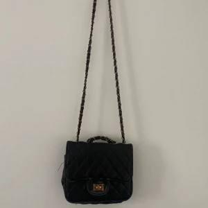 Svart quiltad axelväska - Snygg svart quiltad axelväska med kedjedetaljer på axelremmen. Väskan har en praktisk vridlåsdetalj framtill och en liten topphandtag för extra stil. Perfekt för att ge en elegant touch till din outfit.
