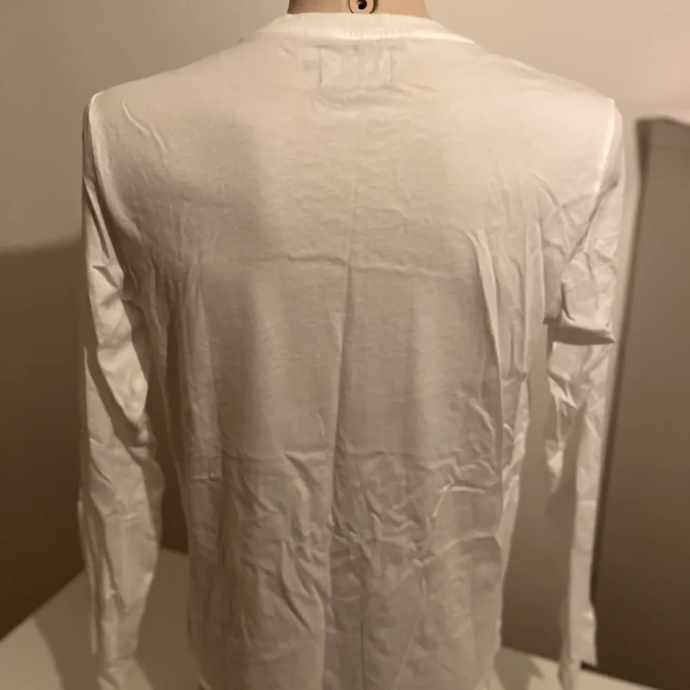 Säljer en stilren vit långärmad tröja från Calvin Klein Jeans med ett stort svart tryck på framsidan. Perfekt för en avslappnad och trendig look. Tröjan är gjord i mjukt bomullsmaterial och har en klassisk passform.. Neuletakit & Villapaidat.