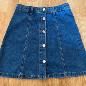 Jeanskjol från H&M Divided - Snygg jeanskjol från H&M Divided i klassisk blå denim. Kjolen har en rad med knappar framtill. Perfekt för en avslappnad stil. Pris går att diskutera. 
