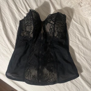 Svart korsett med spetsdetaljer - Elegant svart korsett med vackra spetsdetaljer och en formande passform. Den har en klassisk design med en feminin touch, perfekt för att lyfta vilken outfit som helst. Korsetten är axelbandslös och har en diskret knäppning baktill.