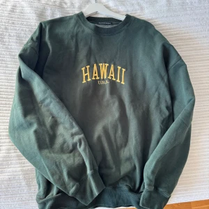 Oversized sweatshirt från Brandy Melville - Säljer en grön sweatshirt från Brandy Melville med texten 'HAWAII U.S.A.' i gult på framsidan. Tröjan har en klassisk rund hals och långa ärmar. Perfekt för en avslappnad stil.