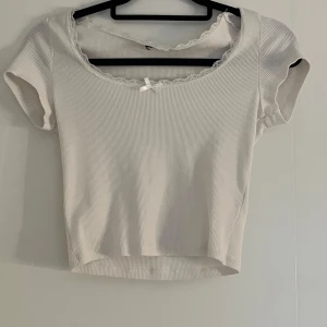 Vit ribbad topp med spetsdetalj - Söt vit ribbad topp med korta ärmar och spetskant vid halsringningen. Toppen har en liten rosett framtill.