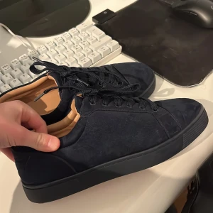 Mörkblå sneakers med röd sula - Snygga mörkblå sneakers med en ikonisk röd sula. Skorna har snörning och är tillverkade i ett elegant material som ger en lyxig känsla. Perfekta för den som vill ha en stilren look med en twist. Bra skick