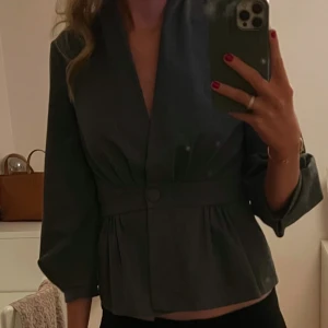 Grå blus med knappar - Snygg grå blus med knappar framtill och trekvartsärmar. Den har en elegant passform med ett brett bälte i midjan som ger en smickrande silhuett. Perfekt för att ge en stilren touch till din outfit. Älskar denna blusen men kommer tyvärr inte till användning längre. 