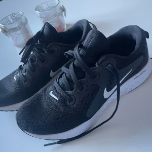 Svarta Nike sneakers - Snygga svarta Nike sneakers med vit sula och klassisk snörning. Perfekta för träning eller vardagsbruk. Skorna har en stilren design med Nike-loggan i vitt på sidan. Strl 38,5, (24,5cm) 