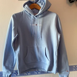 Ljusblå hoodie från Tommy Jeans - Säljer en snygg ljusblå hoodie från Tommy Jeans med broderad logga på bröstet. Hoodien har en klassisk känguruficka och justerbar dragsko i huvan. Perfekt för en avslappnad stil.