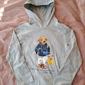 Grå hoodie med Polo Bear från Ralph Lauren - Säljer en grå hoodie från Ralph Lauren med en söt Polo Bear-illustration på framsidan. Tröjan har långa ärmar och är perfekt för en avslappnad stil. Den är tillverkad i mjukt material för extra komfort. Skick 8/10, tveka inte på att ställa frågor , M I BARN STORLEK