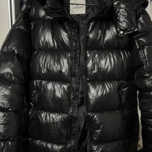 Svart dunjacka från Moncler - Säljer en snygg svart dunjacka från Moncler. Jackan har en glansig finish och är utrustad med en dragkedja framtill. Den har långa ärmar och en praktisk huva. Perfekt för kyliga dagar! Använd bara några gånger 