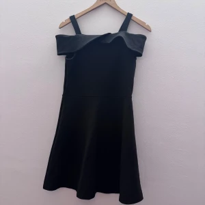 Svart klänning med volangdetalj - Elegant svart klänning med axelbandslös design och volangdetalj vid halsen. Perfekt för en stilren look. Klänningen har en kort längd och är tillverkad i ett mjukt material som ger en bekväm passform.