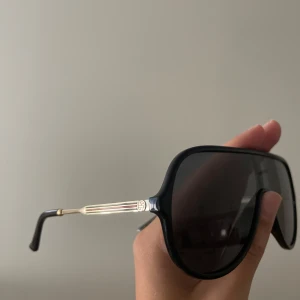 Svarta solglasögon från Gucci - Snygga svarta  solglasögon från Gucci med en klassisk aviatorform. Bågarna har en elegant gulddetalj på sidorna och kommer med ett lyxigt gult fodral. Perfekta för att ge en stilren touch till din look. 