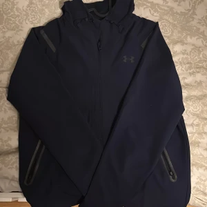 Mörkblå jacka från Under Armour - Säljer en mörkblå jacka från Under Armour med dragkedja och huva. Jackan har långa ärmar och praktiska fickor med dragkedjor. Perfekt för sportiga aktiviteter eller vardagsbruk.