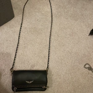 Svart axelväska från Zadig & Voltaire - Snygg svart axelväska i skinn från Zadig & Voltaire med kedjerem och coola nitar. Väskan har en dragkedja och ett stilrent emblem med vingar på framsidan. Perfekt för att ge din outfit en edgy touch! På de två sista bilderna ser man att den tappat lite färg men syns bara på nära håll! Därav priset , ny pris 2500.