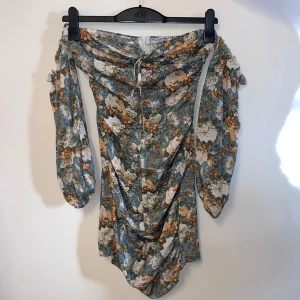 Blommig klänning från Glamaker - Säljer en superfin blommig klänning från Glamaker. Den har en offshoulder-design med långa ärmar och ett vackert blommönster i blått, vitt och orange. Perfekt för en somrig look! 💐