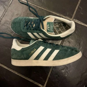 Adidas Gazelle i grön mocka - Snygga Adidas Gazelle sneakers i grön mocka med vita detaljer och snörning. Klassisk design med tre vita ränder på sidan och Gazelle-logga i guld. Perfekta för en stilren look.