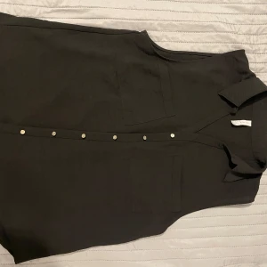 Svart ärmlös skjorta från Amisu - Snygg svart ärmlös skjorta från Amisu med knappar framtill. Perfekt för en stilren look. Skjortan har en klassisk krage och två bröstfickor som ger en extra touch.