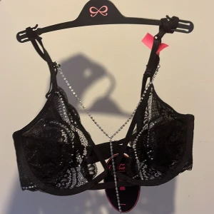Svart spetskorsett med strassdetaljer - Säljer en elegant svart spetskorsett från Victoria's Secret med vackra strassdetaljer och korsade band framtill. Helt nya med tagg, storlek 70S samt underdel storlek S