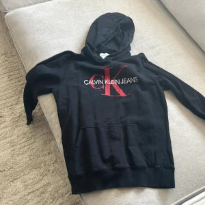 Svart hoodie från Calvin Klein Jeans - Säljer en stilren svart hoodie från Calvin Klein Jeans med röd och vit logga på framsidan. Tröjan har en klassisk känguruficka och en bekväm huva. Perfekt för en avslappnad look.