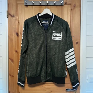 Grön djurmönstrad bomberjacka från Svea - Snygg grön bomberjacka från Svea med vita detaljer och dragkedja. Jackan har ett diskret mönster och vita ränder på ärmarna. Perfekt för en cool och avslappnad stil.