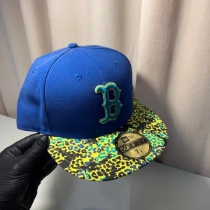 New Era keps - Snygg blå keps från New Era med ett iögonfallande leopardmönster i gult och blått på skärmen. Kepsen har en broderad bokstav 'B' framtill och logotypdetaljer på sidan och baksidan. Perfekt för att sticka ut i mängden!