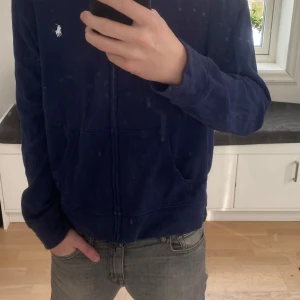 Mörkblå hoodie från Ralph Lauren - Säljer en snygg mörkblå hoodie från Ralph Lauren med dragkedja och klassisk logga på bröstet i storlek 164. Perfekt för en avslappnad stil. Den har långa ärmar och en bekväm passform.