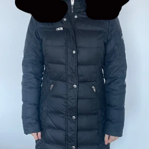 Svart dunjacka från Rockandblue - Snygg svart dunjacka från Rockandblue med quiltad design och dragkedjor. Jackan har en lång passform och är perfekt för kyliga dagar. Märkeslogga på ärmen ger en extra touch. Perfekt för dig som vill hålla stilen i vinter! ❤️